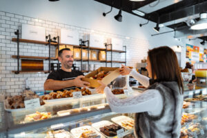 A smiling male bakery worker hands a box of pastries to a female customer across a glass display counter.
