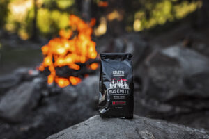 Black coffee bag labeled Yosemite whole bean sits on rock with campfire burning behind.