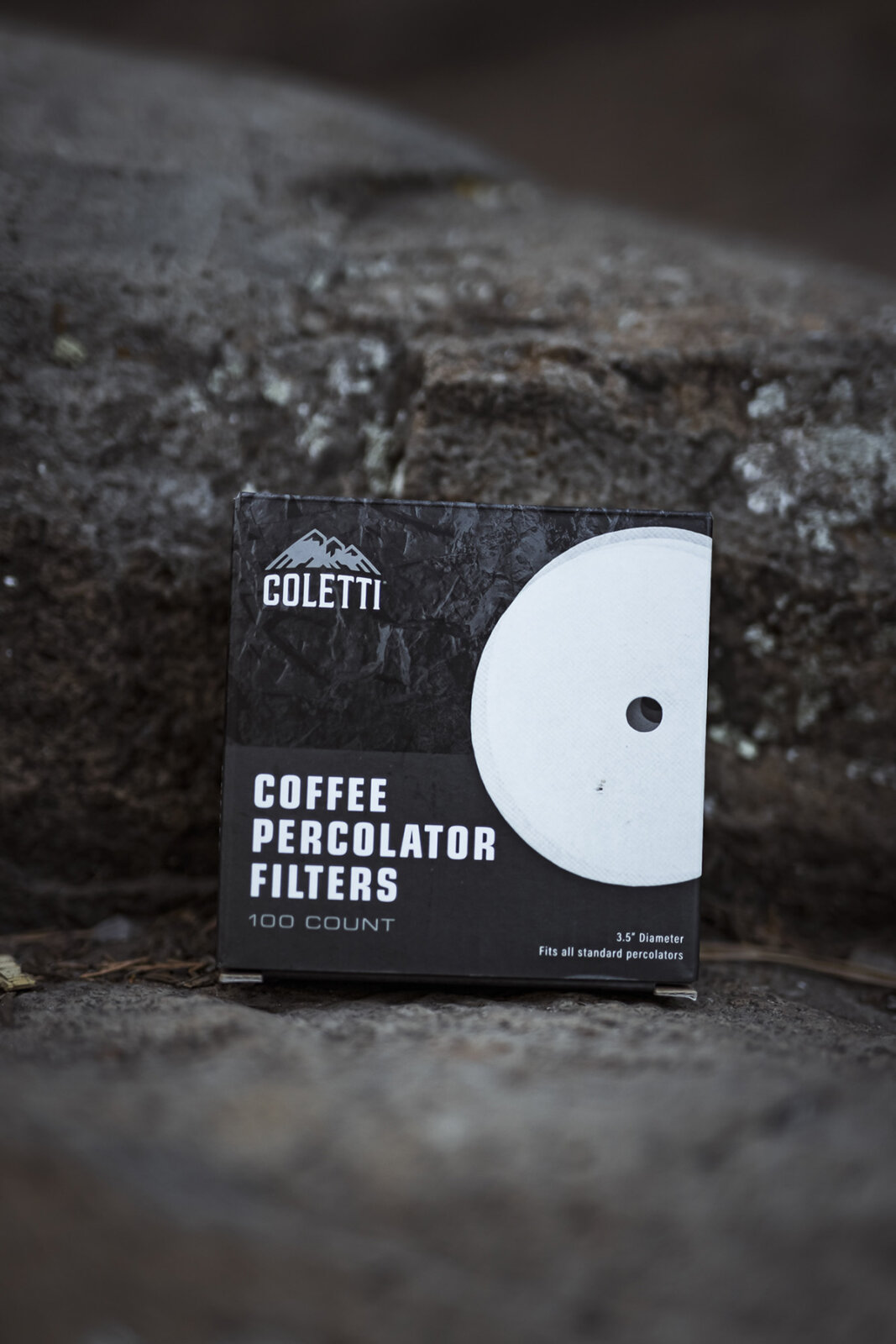 This product photograph features a black box of coffee percolator filters positioned on a rough, weathered stone surface. The packaging displays a white circular filter disc with a center hole. Soft, natural lighting creates a shallow depth of field, blurring the stone background while keeping the product sharp and prominent.