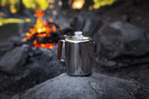 Stainless steel coffee percolator with wooden handle sits on rock near campfire.