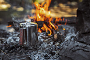 Stainless steel coffee percolator with wooden handle sits on grill grate beside crackling campfire.