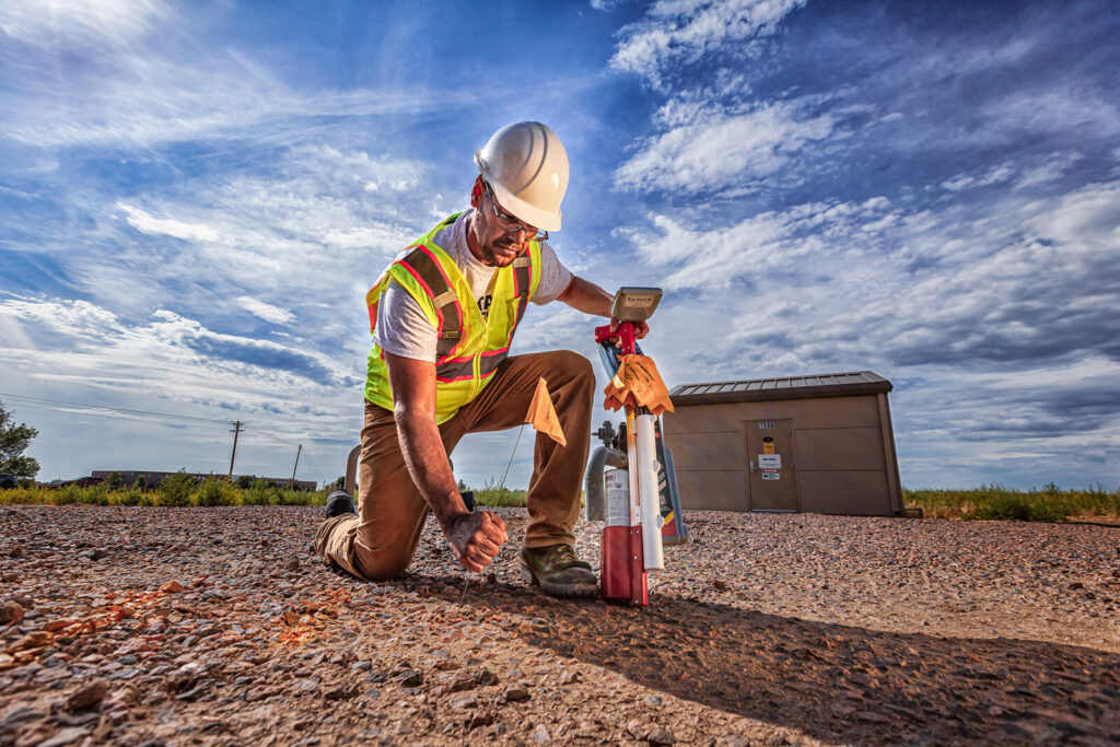 Construction worker in safety vest kneeling on gravel using survey equipment under dramatic clouds.