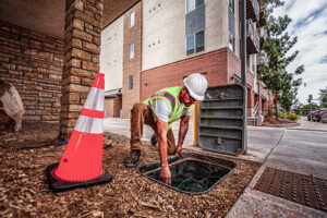 Utility worker in safety vest and hard hat inspects underground cable box near sidewalk.