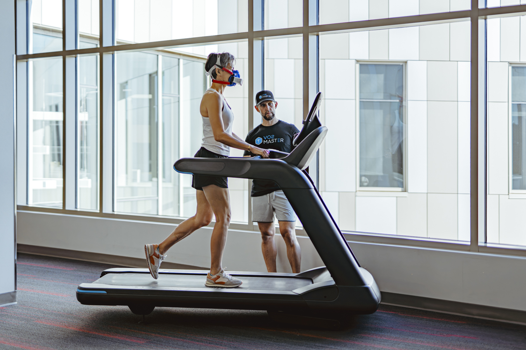 Female athlete walking on treadmill wearing VO2 Master metabolic analyzer during indoor performance test with trainer observing