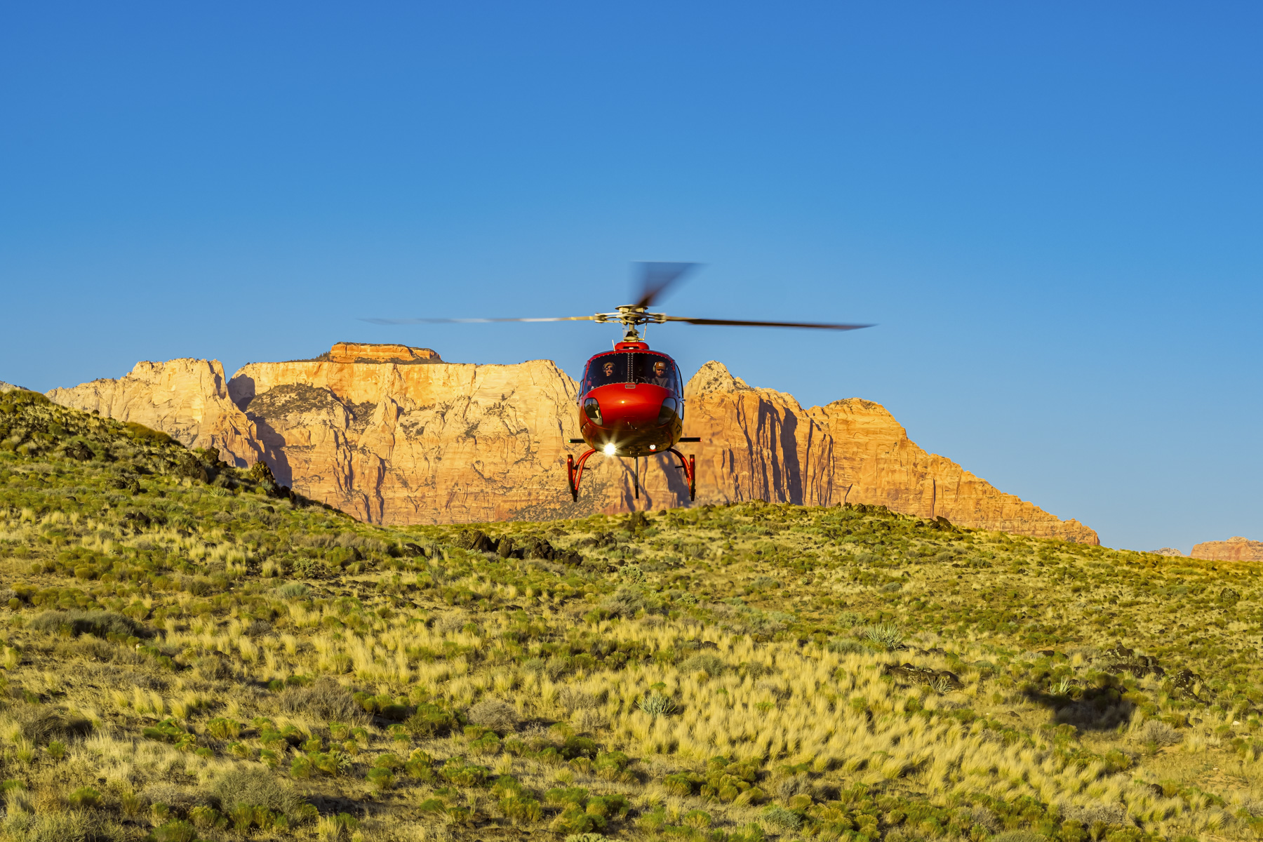 Red helicopter with spinning rotors landed on green hillside with golden Zion cliffs captured through professional commercial photography