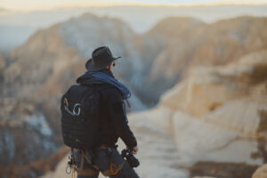Photographer in full hiking gear stands on cliff edge at sunset in Southern Utah holding camera with rope on backpack