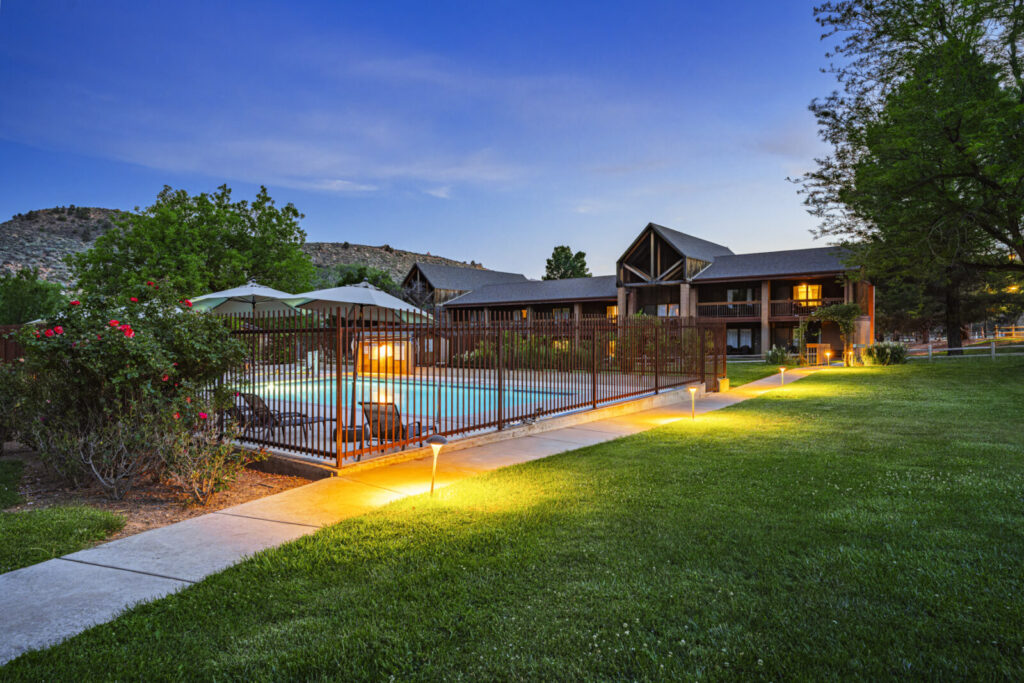 A fenced outdoor pool glows with warm lights beside a rustic multi-story lodge at dusk.