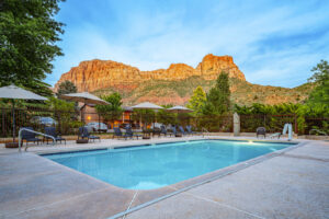 A rectangular swimming pool with lounge chairs and umbrellas set against towering red rock formations.