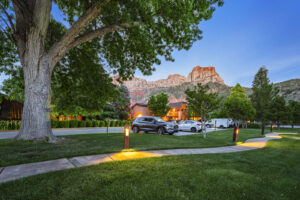 A landscaped lawn with glowing path lights, parked cars, and red rock formations in the background at dusk.