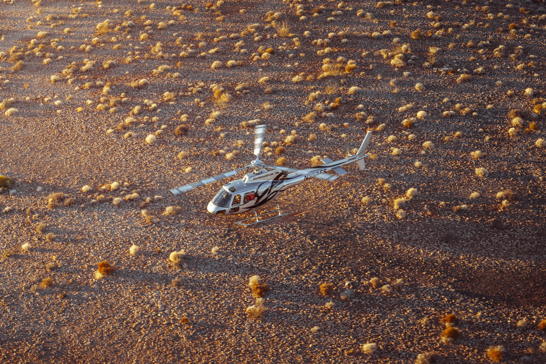 Helicopter hovers above desert brush and rocky plateau in warm afternoon light, photographed for aviation tourism and travel brand marketing.