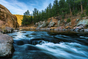 Fast-moving whitewater cascades over smooth rocks through a narrow forested canyon with pine trees and rocky cliffs.