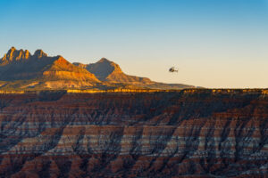 Helicopter flies over striped red and gray badlands with sunlit rugged mountain peaks in background.