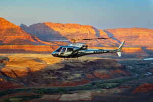 A helicopter flies over dramatic red rock formations bathed in warm golden sunset light.