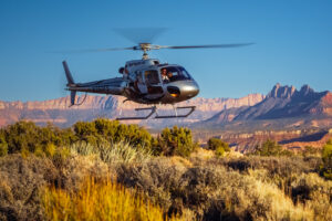 Dark helicopter hovering low over desert brush with red rock mountain formations in background.