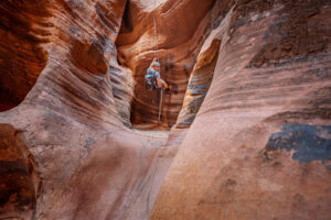 A canyoneer rappels down a rope through a narrow slot canyon with sculpted red sandstone walls.