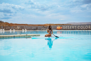 A woman in a floral swimsuit sits on a white paddleboard holding a paddle on calm blue water.