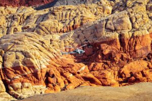 A helicopter flies past dramatic red and yellow layered rock formations in rugged terrain.