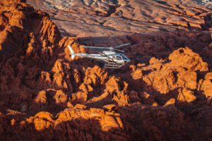 White helicopter flies low over rugged red and orange rocky terrain in warm golden light.