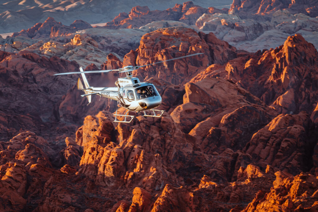 Silver helicopter flies low over dramatic red and orange rocky terrain in warm golden light.