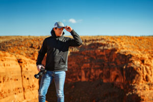 A photographer holding a camera adjusts his cap while standing near a deep red rock formation under a clear blue sky.