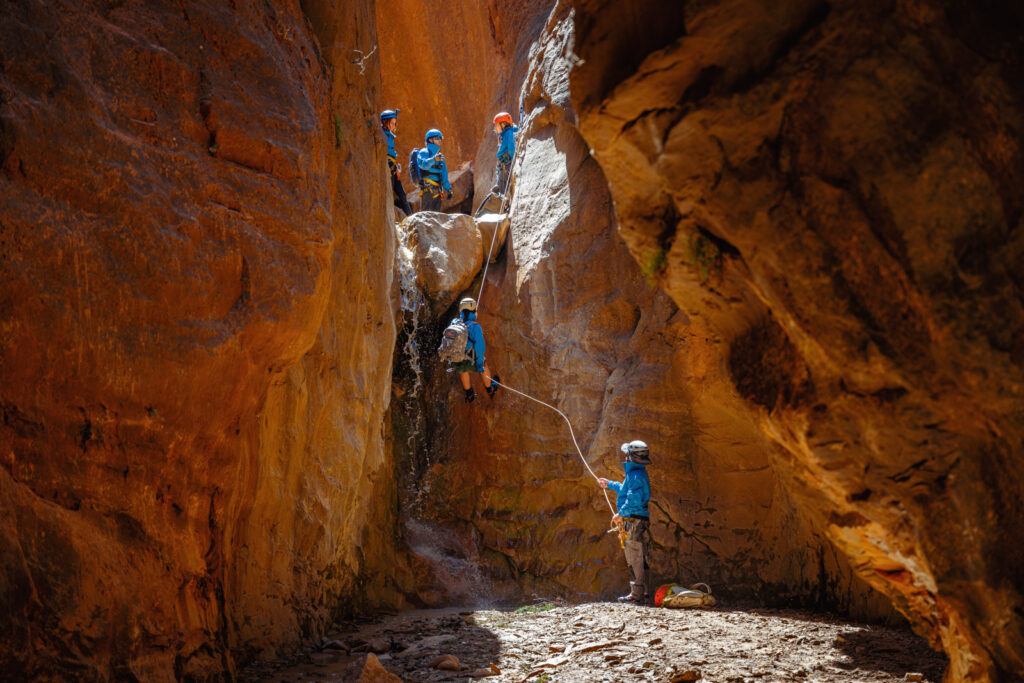 Group of canyoneers in blue shirts and helmets rappelling through a narrow red rock slot canyon.