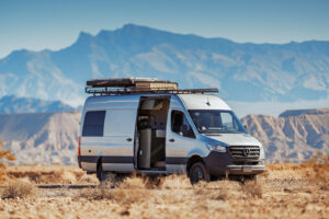 A silver converted cargo van with a roof rack sits on dry rocky terrain with rugged mountains behind it.