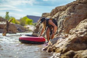 A person in a bikini prepares a red and black inflatable tube in shallow water near rocky terrain.
