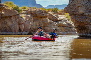Two people floating on inflatable tubes in brown river water between rocky canyon walls.