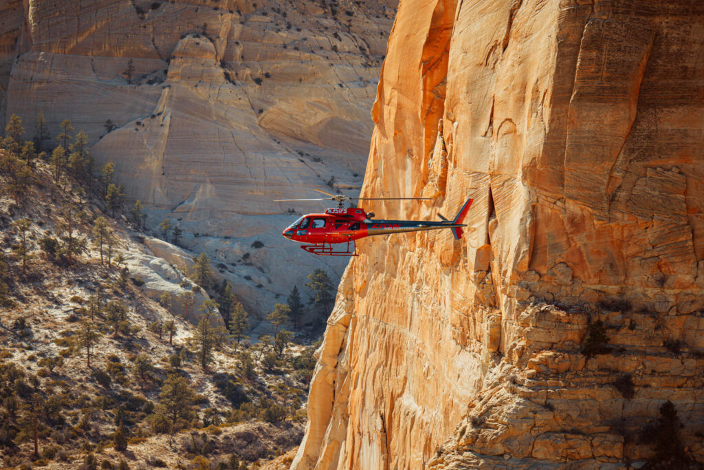 Red helicopter flies near massive layered sandstone cliff walls in a rugged canyon landscape.