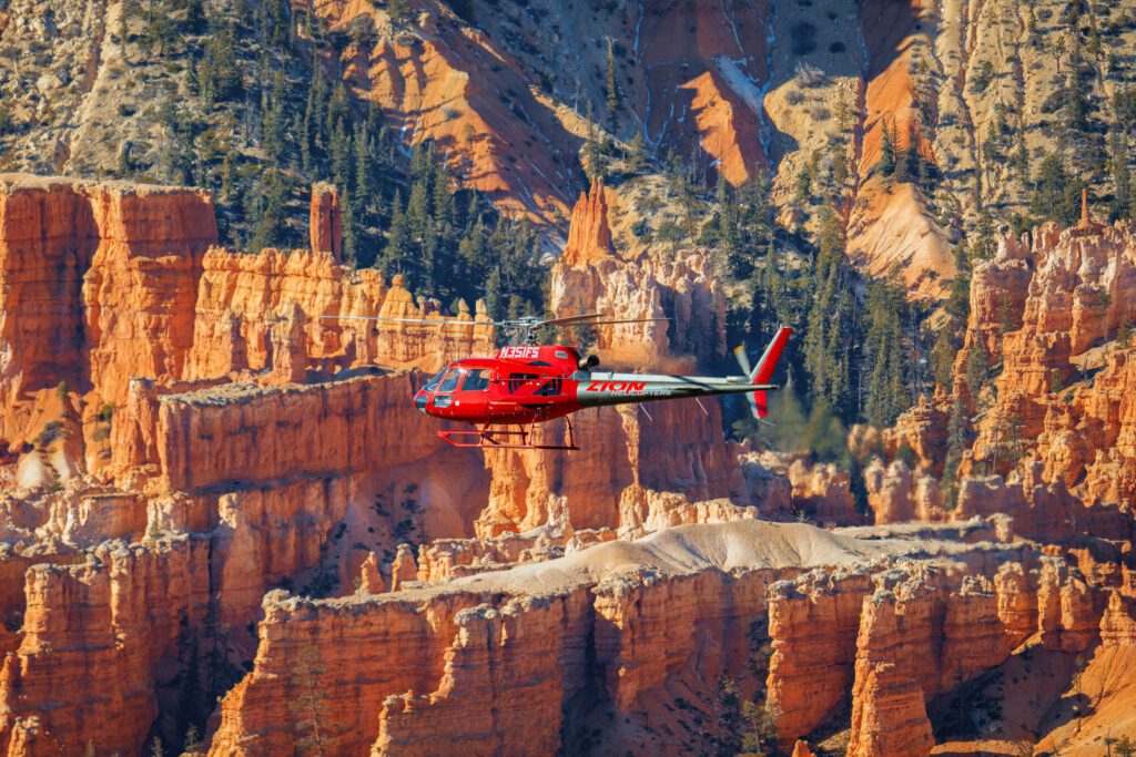 A red helicopter flies in front of tall, layered orange and red rock spires with scattered evergreen trees.