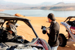 A photographer holds a camera toward a helmeted rider seated inside an off-road side-by-side vehicle on sandy terrain.
