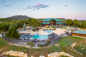 Aerial view of a resort lodge with a turquoise roof and two swimming pools with gazebos, surrounded by evergreen trees and mountains at dusk.