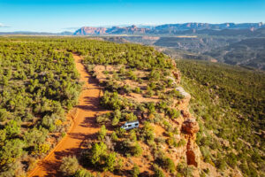 An aerial view of a white recreational vehicle parked along a red dirt road near a rocky cliff edge surrounded by green shrubland.