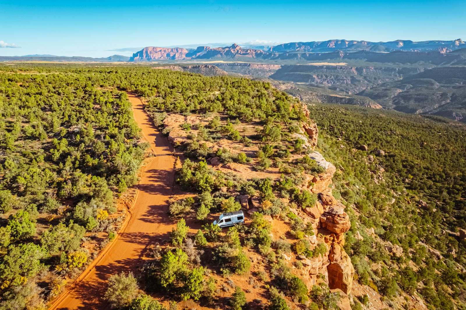 An aerial view of a white recreational vehicle parked along a red dirt road near a rocky cliff edge surrounded by green shrubland.