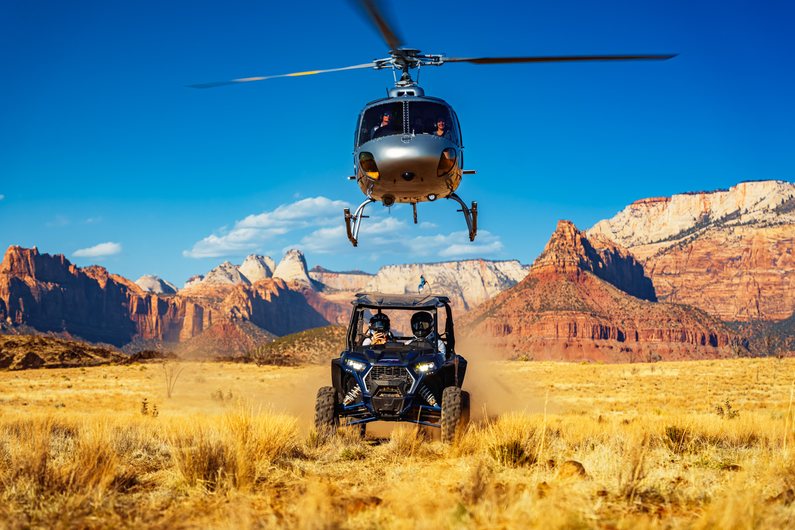 Helicopter flying above an off-road vehicle among red rock formations and golden grassland