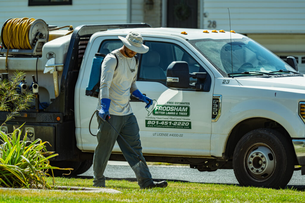 A lawn care technician carries a backpack sprayer while walking beside a white service truck.