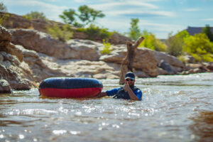 A person in blue shirt swimming while pulling a red and blue inflatable tube through clear water.