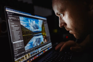 A person leans close to a laptop screen displaying a landscape photo editing application in a dark room.
