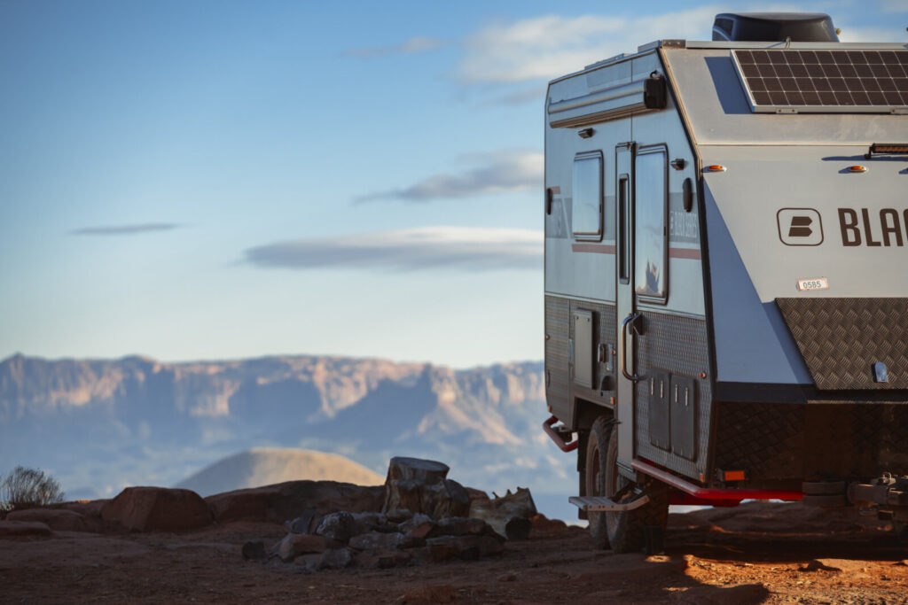 A rugged travel trailer with rooftop solar panels parked on rocky terrain with layered mountains in the background.