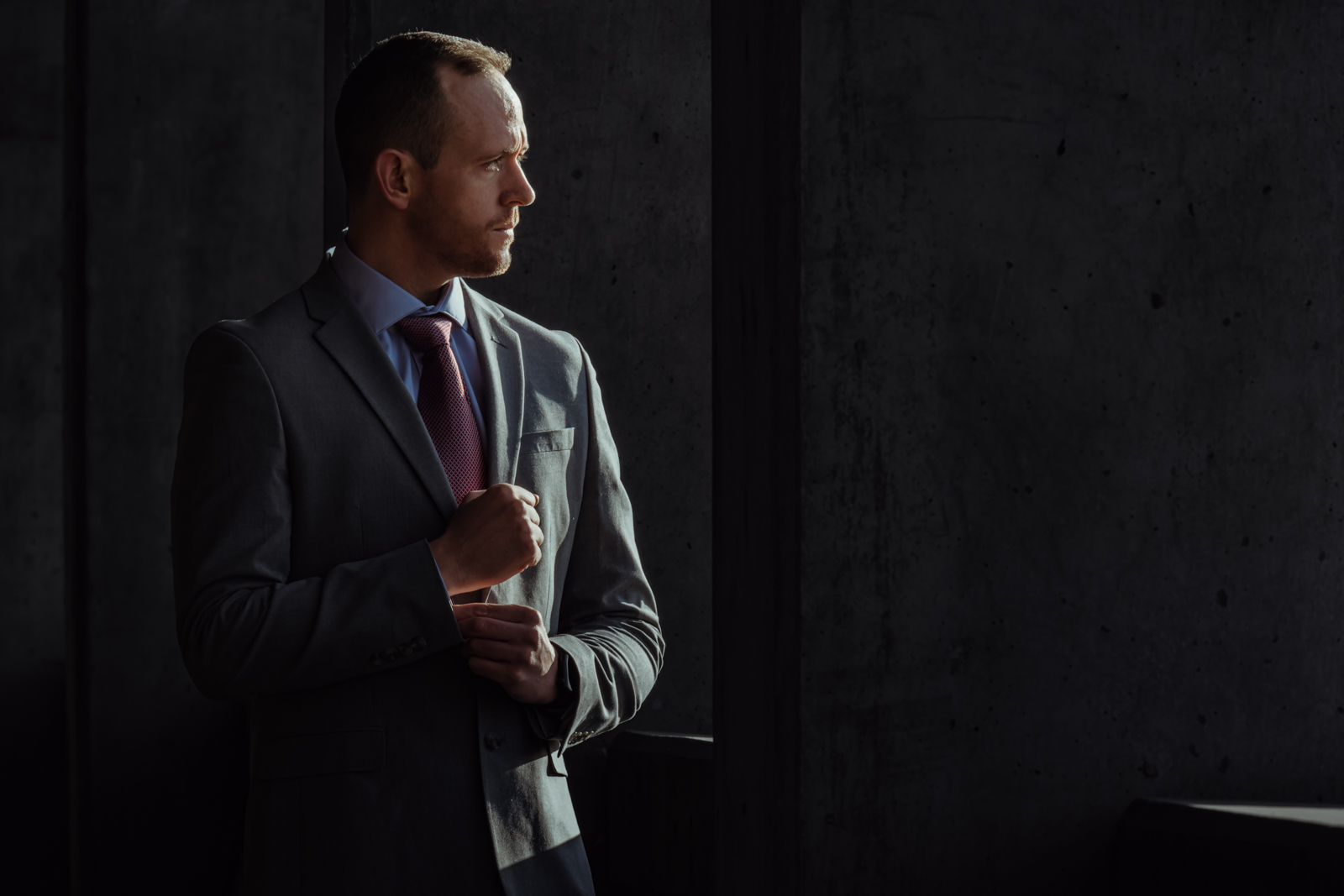 A man in a gray suit and burgundy tie stands near a window, adjusting his jacket lapel.