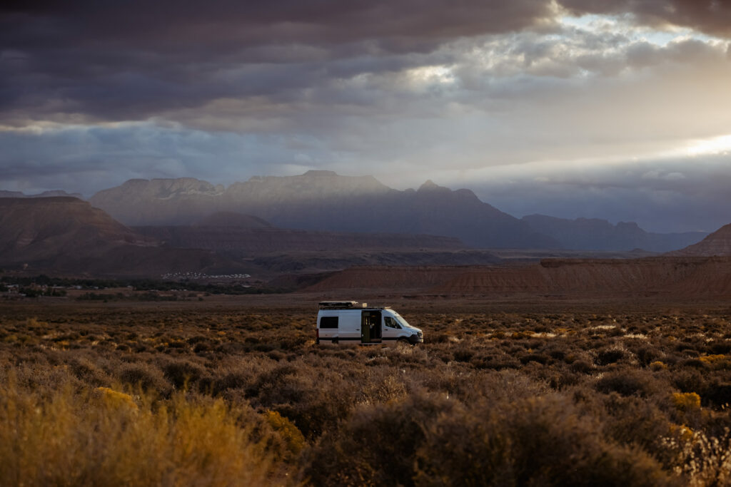 A white camper van sits alone in a wide, flat scrubland with mountains and stormy clouds behind it.