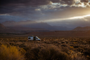 A white camper van sits alone in a wide open scrubland landscape beneath a stormy, cloud-filled sky.