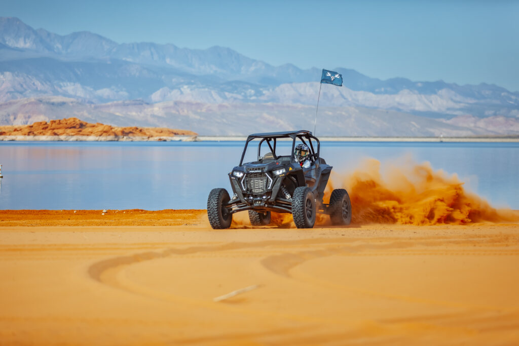 Mad Moose Rentals side-by-side kicking up sand on the beach at Sand Hollow Reservoir with a branded flag and mountains in the background.